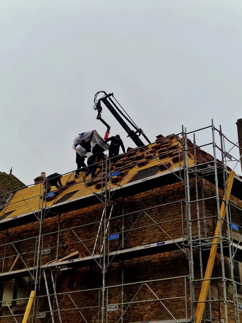 Deux couvreurs sur une toiture en tuiles anciennes, grue en soutien, échafaudage sur toute la hauteur de la façade en pierre