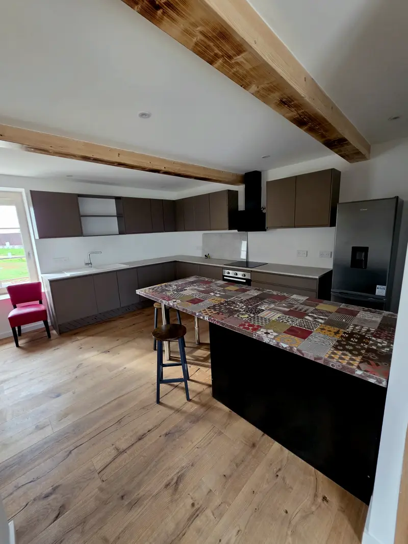 Cuisine livrée sous poutres apparentes, îlot coiffé de carreaux de ciment dépareillés, parquet chêne